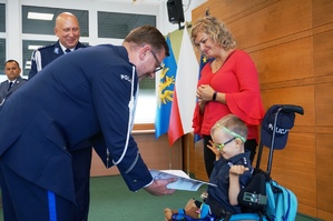 grafika-sala, dziecko na wózku inwalidzkim, komendant Łączyk wręcza akt mianowania Szymkowi