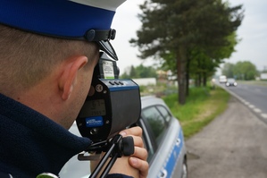 grafika, dzień, droga, pochmurno, policjant patrzy przez urządzenie do pomiaru prędkości na samochody