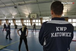 grafika-sala gimnastyczna, mężczyzna w czarnej koszulce z napisem KWP Katowice, w tle ćwiczą osoby