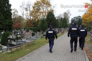 grafika-dzień, trójka policjantów spaceruje po rejonie cmentarza, widoczne nagrobki