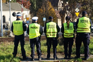 dzień, słonecznie, funkcjonariusze stoją odwróceni plecami i obserwują pieszych na przejściu kolejowym