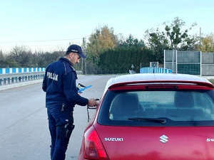 grafika - dzień ,pochmurno, policjant stoi przy drzwiach kierowcy i wręcza ulotki oraz bada stan trzeźwości