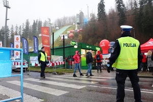grafika-dzień, droga, policjanci na drodze i przejście dla pieszych, w tle skocznia i radiowozy oraz kibice