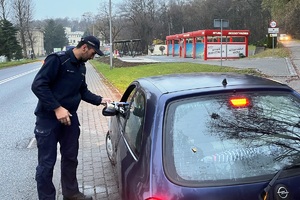 grafika-dzień, pochmurno, policjant przy aucie od strony kierowcy wręcza kierującemu gadżety akcji