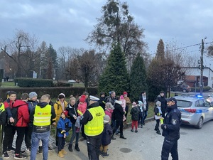 zdjęcie-dzień, policjanci rozdają odblaski pieszym na parkingu, widoczne wozy policyjne i straży pożarnej z włączonymi niebieskimi sygnałami uprzywilejowania