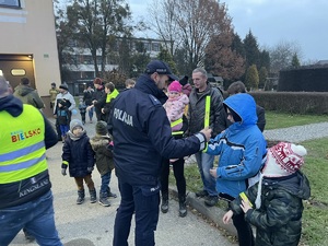 zdjęcie-dzień, policjanci rozdają odblaski pieszym na parkingu, widoczne wozy policyjne i straży pożarnej z włączonymi niebieskimi sygnałami uprzywilejowania