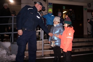 grafika- zima, noc, oświetlenie sztuczne, w tle budynek, Zdjęcie przedstawia policjanta odbierającego świeczkę od dzieci