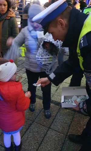 zdjęcie-noc, sztuczne światło, policjant schyla się i wręcza element odblaskowy dziewczynce