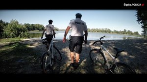 grafika-dzień, słonecznie, policjanci przy rowerach, w tle jezioro