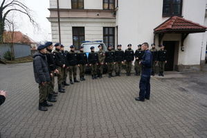 zdjęcie- młodzież z klasy mundurowej stoi na placu parkingowym przy komendzie i słucha policjanta