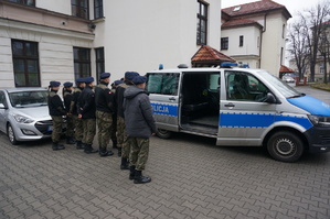 zdjęcie- młodzież z klasy mundurowej stoi na placu parkingowym przy komendzie i słucha policjanta