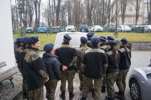 zdjęcie- młodzież z klasy mundurowej stoi na placu parkingowym przy komendzie i słucha policjanta