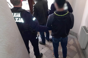 zdjęcie-pomieszczenie, korytarz, policjant pilnuje zatrzymanego, który ma kajdanki na rękach trzymanych z tyłu