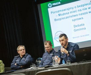 grafika-pomieszczenie, sztuczne oświetlenie, policjanci siedzą za stołem, rozmawiają z zebranymi w sali ludźmi