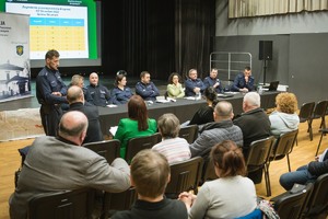 grafika-pomieszczenie, sztuczne oświetlenie, policjanci siedzą za stołem, rozmawiają z zebranymi w sali ludźmi