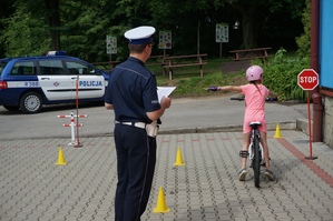grafika-policjant obserwuje jak dziecko jedzie na rowerze, plac, znaki drogowe, dzień