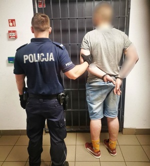 zdjęcie-pomieszczenie, policjant trzyma mężczyznę za łokieć, mężczyzna ma kajdanki na rękach zapięte z tyłu ,