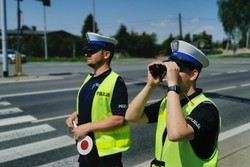 zdjęcie, dzień słonecznie, policjant przez lornetkę obserwuje ruch samochodów przy drodze