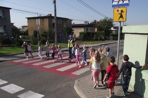 zdjęcie-dzieci przechodzą przez jezdnię, na pasach stoi policjantka i ich obserwuje, słonecznie