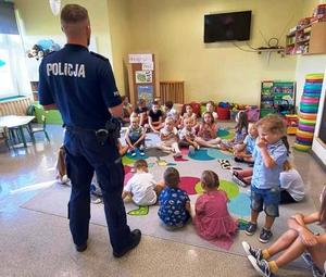 zdjęcie-policjant stoi i przemawia, przedszkolaki siedzą na dywanie i słuchają