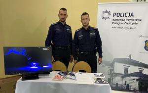 zdjęcie-policjanci obok siebie, stoją przez stołem z monitorem i ulotkami, za nimi baner z napisem Komenda Powiatowa Policji w Cieszynie