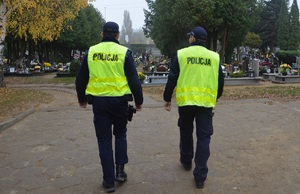 zdjęcie-dwóch policjantów w zielonych odblaskowych kamizelkach z napisem POLICJA patrolują cmentarz