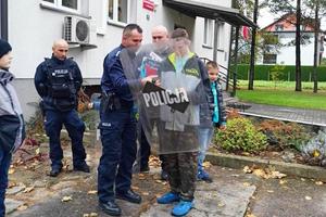 zdjęcie na placu przed komisariatem. dzień ,pochmurnie, policjant podaje dziecku tarczę z napisem policja, inne osoby się przygladają