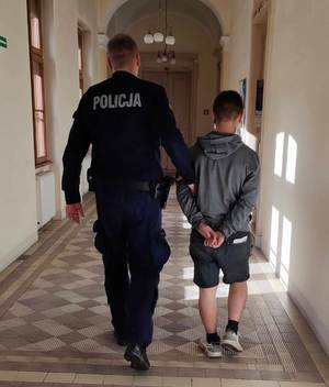 zdjęcie-korytarz, policjant prowadzi za ramię zatrzymanego mężczyznę, który ma kajdanki na rękach zapięte z tyłu