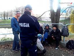 policjant rozmawia z siedzącymi ludźmi, dzień, w tle rzeka