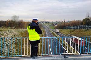 zdjęcie-dzień, pochmurno, policjant patrzy przez miernik prędkości na drogę, sprawdza prędkość samochodu i odległość między pojazdami