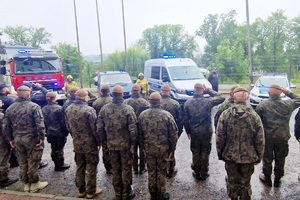 zdjęcie- dzień,pochmurno, służby mundurowe stoją na placu w pozycji zasadniczej, oddają honory, w tle radiowozy