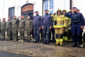 zdjęcie- dzień,pochmurno, służby mundurowe stoją na placu w pozycji zasadniczej, oddają honory, w tle radiowozy