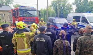 zdjęcie- dzień,pochmurno, służby mundurowe stoją na placu w pozycji zasadniczej, oddają honory, w tle radiowozy