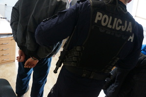 policjant trzyma za łokieć osobę z kajdankami na rękach, pomieszczenie