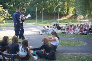 zdjęcie- dzień, słonecznie, dzieci siedzą na trawie w parku, słuchają przemawiających policjantów