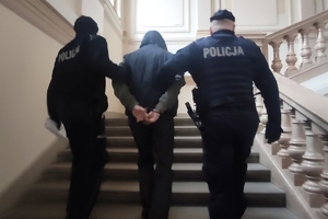 zdjęcie-policjanci prowadzą mężczyznę z kajdankami na rękach z tyłu, idą po schodach w górę w budynku sądu