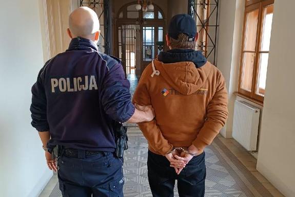 zdjęcie-policjant trzyma za łokieć zatrzymanego, który ma kajdanki na rękach z tyłu. Stoją w korytarzu, dzień