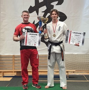 Szymon Bloch z dyplomem i pucharem, ubrany w karate-gi, obok Komendant i trener w jednym Arkadiusz Bloch