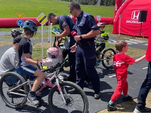dzień, pogoda słoneczna, policjanci i dzieci w miasteczku ruchu, dzieci na rowerach, hulajnogach, rozmawiają z policjantami