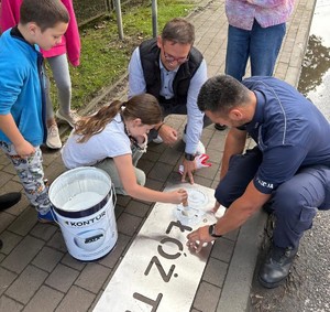 dzień, droga, policjant z dziećmi maluje napis przed przejściem dla pieszych