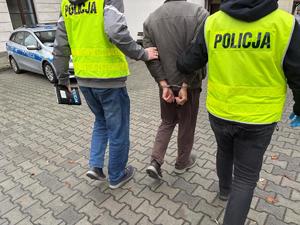 dzień, policjanci prowadza zatrzymanego mężczyznę, ma kajdanki na rękach z tyłu