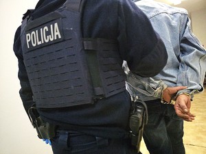 policjant prowadzi osobę korytarzem, zatrzymany ma kajdanki na rekach z tyłu, oświetlenie sztuczne