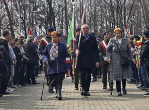 Dzień, słonecznie, plac przed pomnikiem, ludzie zebrani na uroczystości - przedstawiciele służb mundurowych, uczniowie szkół, mieszkańcy, widoczne flagi Polski oraz sztandary