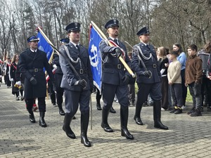 Dzień, słonecznie, plac przed pomnikiem, ludzie zebrani na uroczystości - przedstawiciele służb mundurowych, uczniowie szkół, mieszkańcy, widoczne flagi Polski oraz sztandary