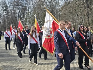 Dzień, słonecznie, plac przed pomnikiem, ludzie zebrani na uroczystości - przedstawiciele służb mundurowych, uczniowie szkół, mieszkańcy, widoczne flagi Polski oraz sztandary