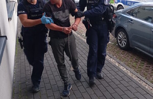 policjanci prowadzą osobę zatrzymana, która ma kajdanki na rekach i nogach, dzień, pochmurno