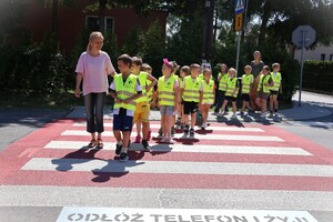 dzień, słonecznie, dzieci przechodzą pod opieką dorosłego przez przejście dla pieszych, widoczny na jezdni przy pasach napis „Odłóż telefon i żyj!”