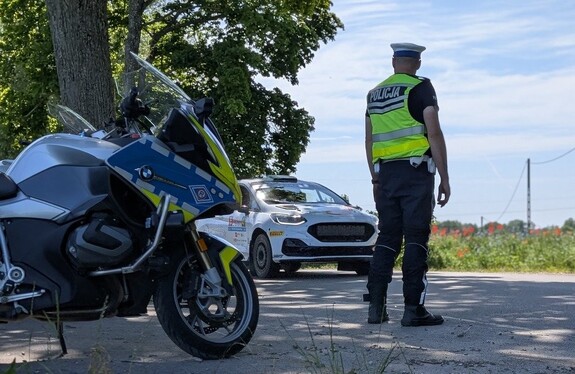 policjant stoi przy motocyklu , w tle droga i samochód rajdowy, dzień